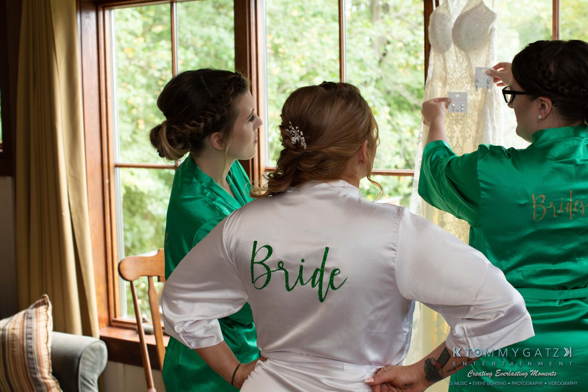 Three women in green and white robes labeled "Bride" look at a hanging white lace dress in a room with large windows and wooden trim.