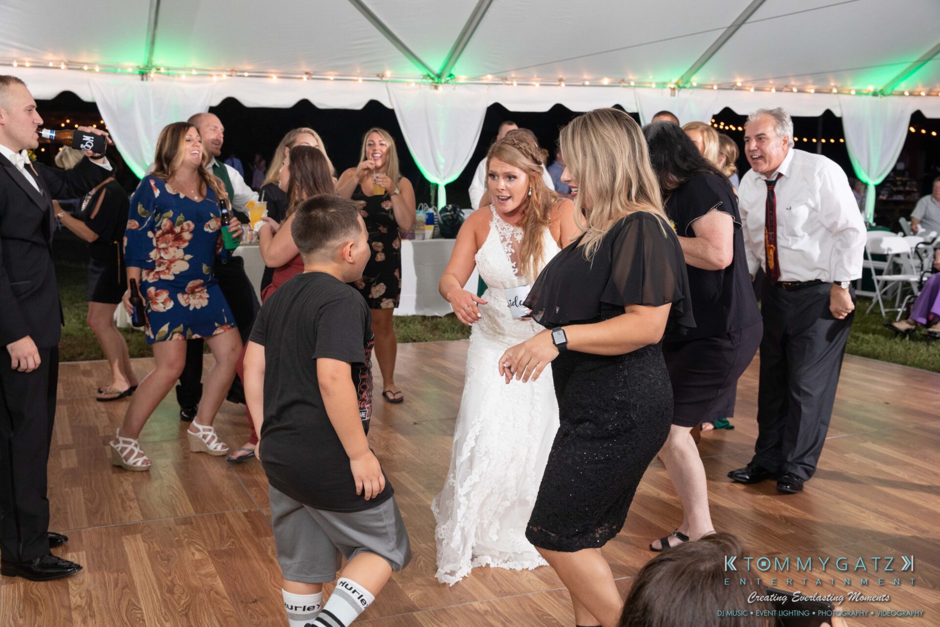 People dancing enthusiastically on a wooden dance floor under a white tent with string lights at a wedding reception.