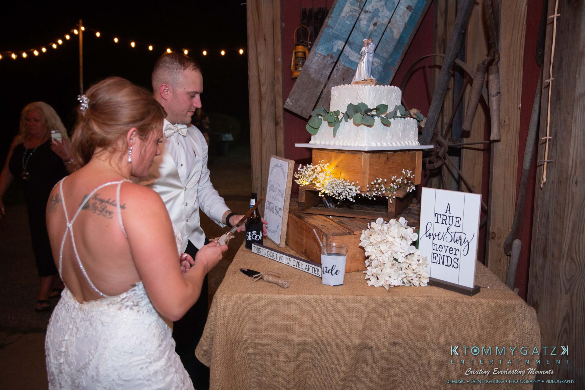 A bride and groom prepare to cut a tiered wedding cake adorned with flowers. A sign reads "A true love story never ends." String lights and a wooden backdrop are in the background.