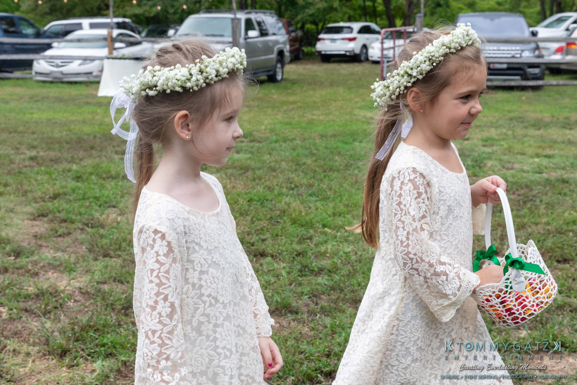 Two young girls in white lace dresses and flower crowns walk on grass. One holds a white basket with green ribbons. Cars and trees are in the background.
