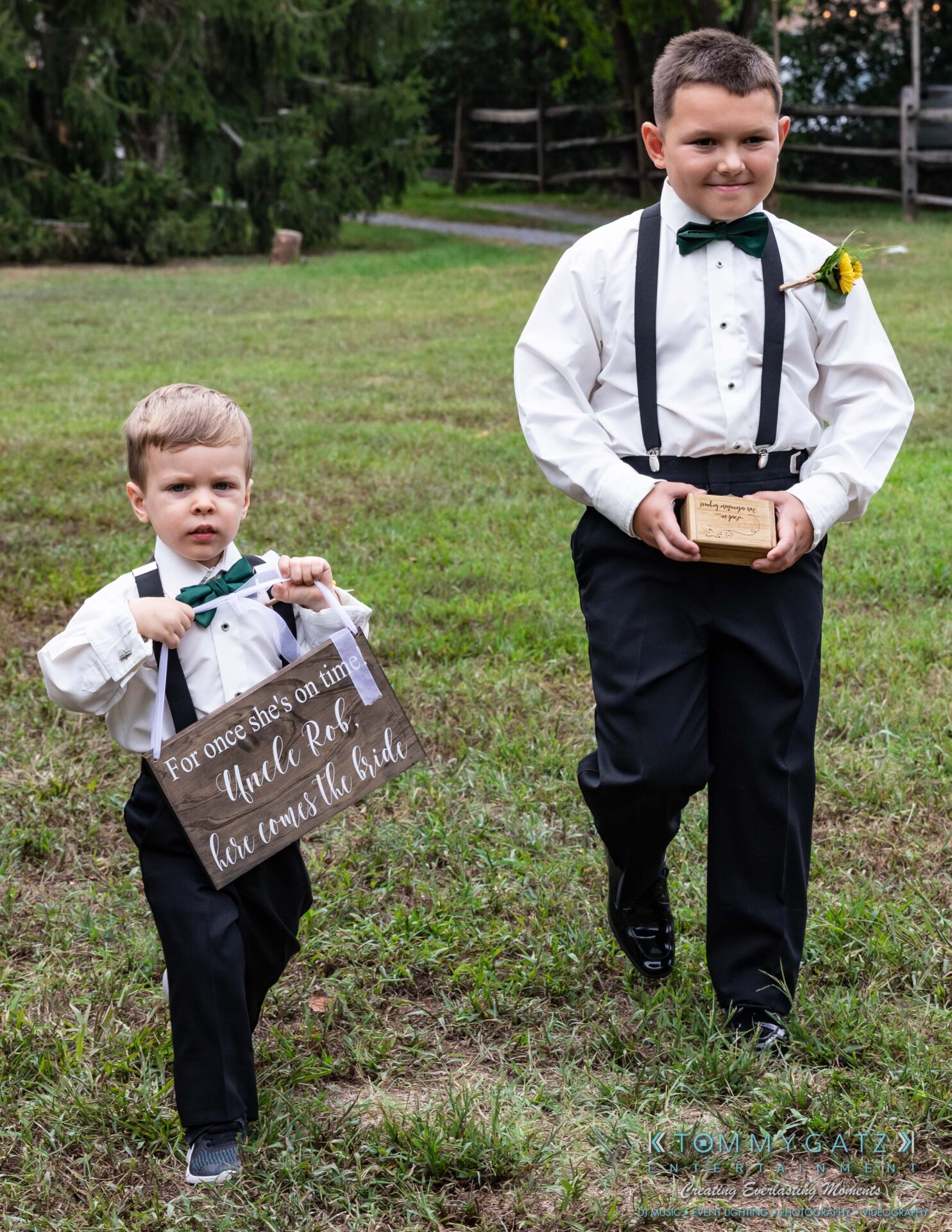 Two young boys dressed in formal attire walk on grass. One holds a sign saying "For once she's on time, Uncle Rob, here comes the bride." The other carries a wooden box.
