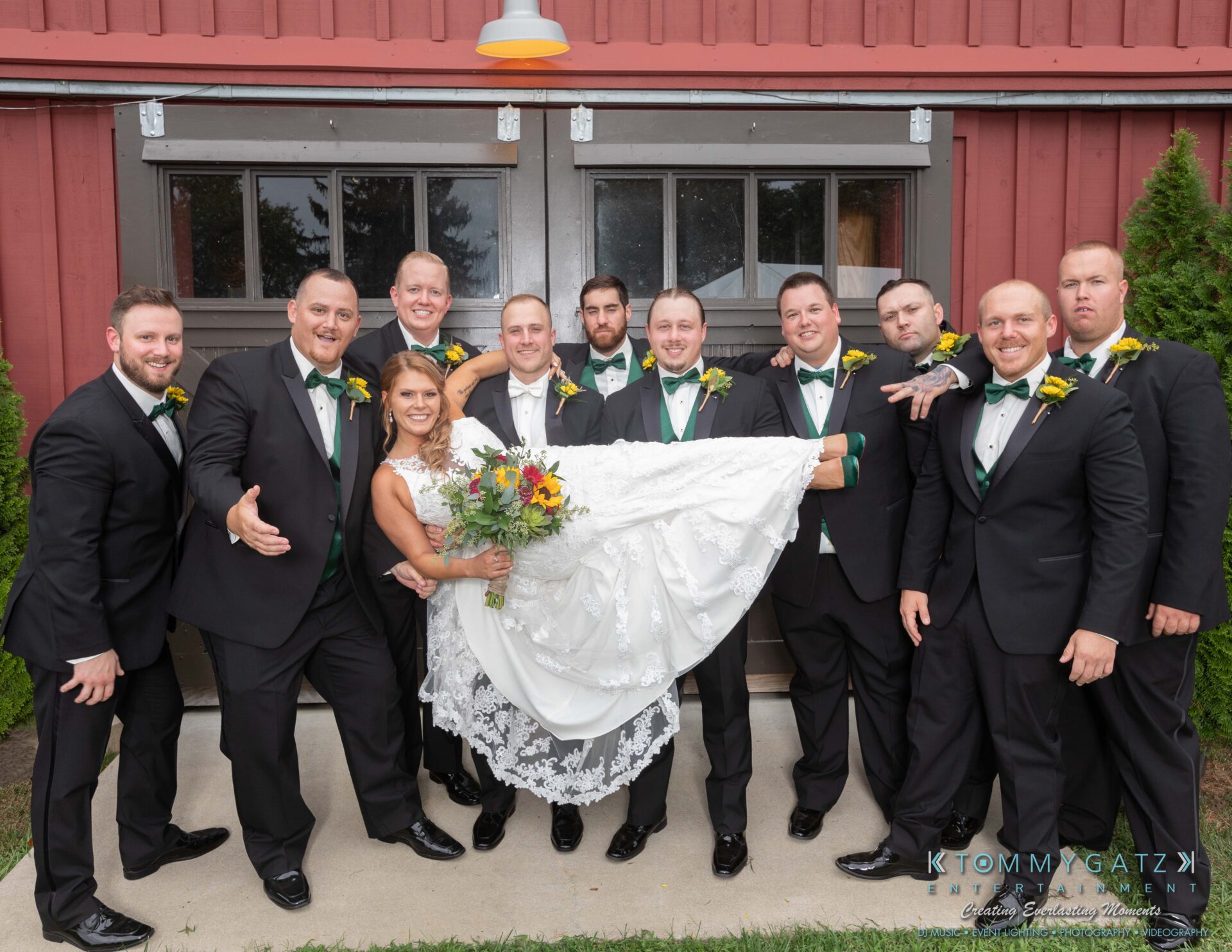 A bride in a white dress is playfully lifted by groomsmen in black suits and green vests in front of a rustic building.