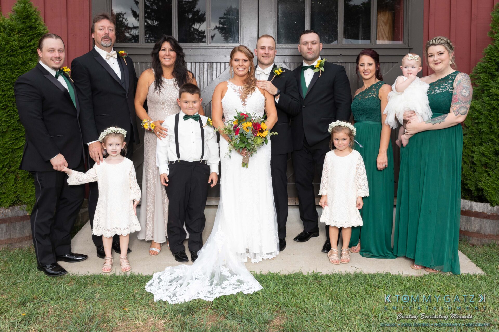 A wedding party poses for a group photo outdoors. The bride and groom stand center, surrounded by flower girls, a ring bearer, bridesmaids, and groomsmen.
