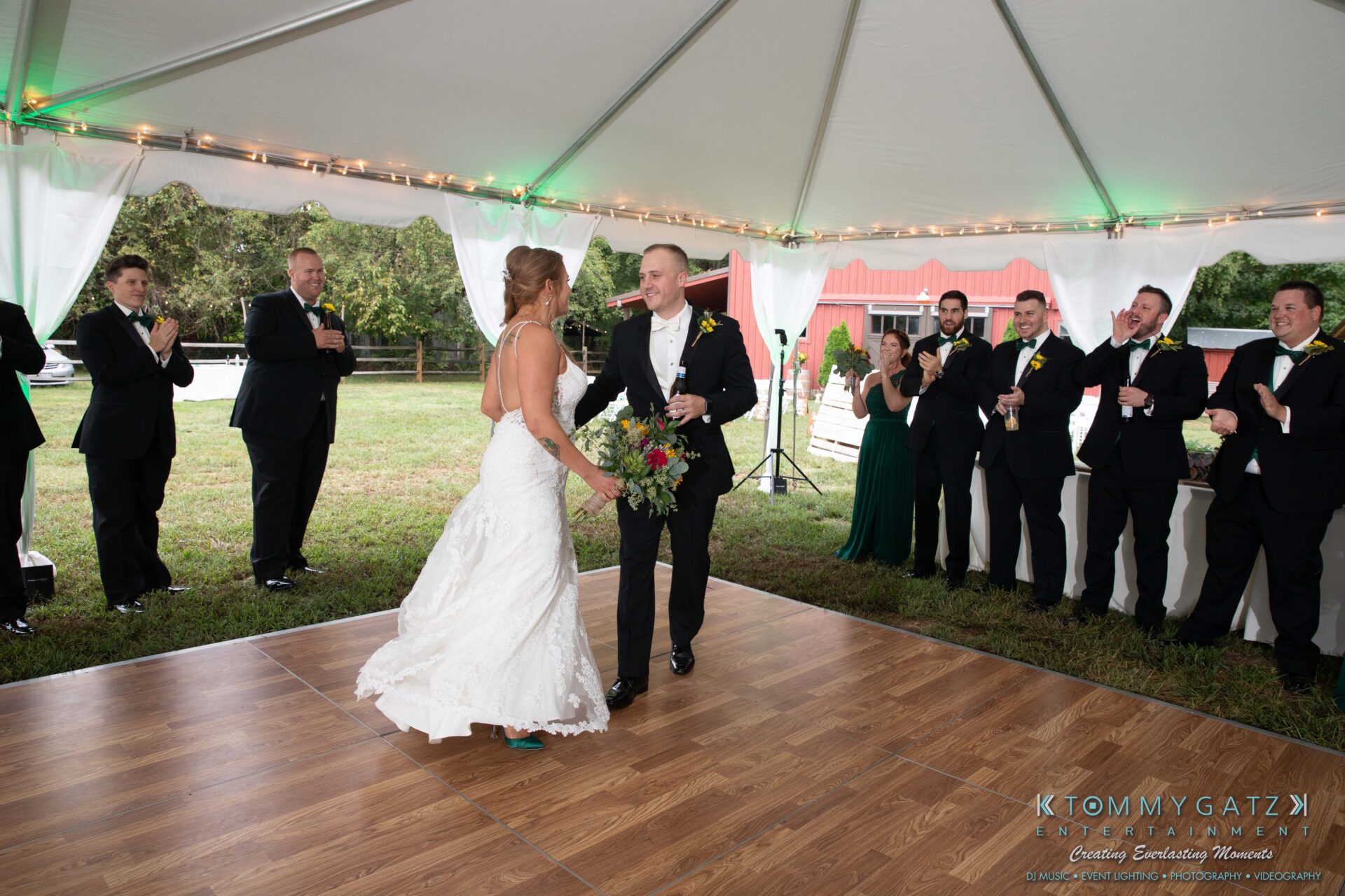 A bride and groom dance under a tent while guests in formal attire stand around and clap.