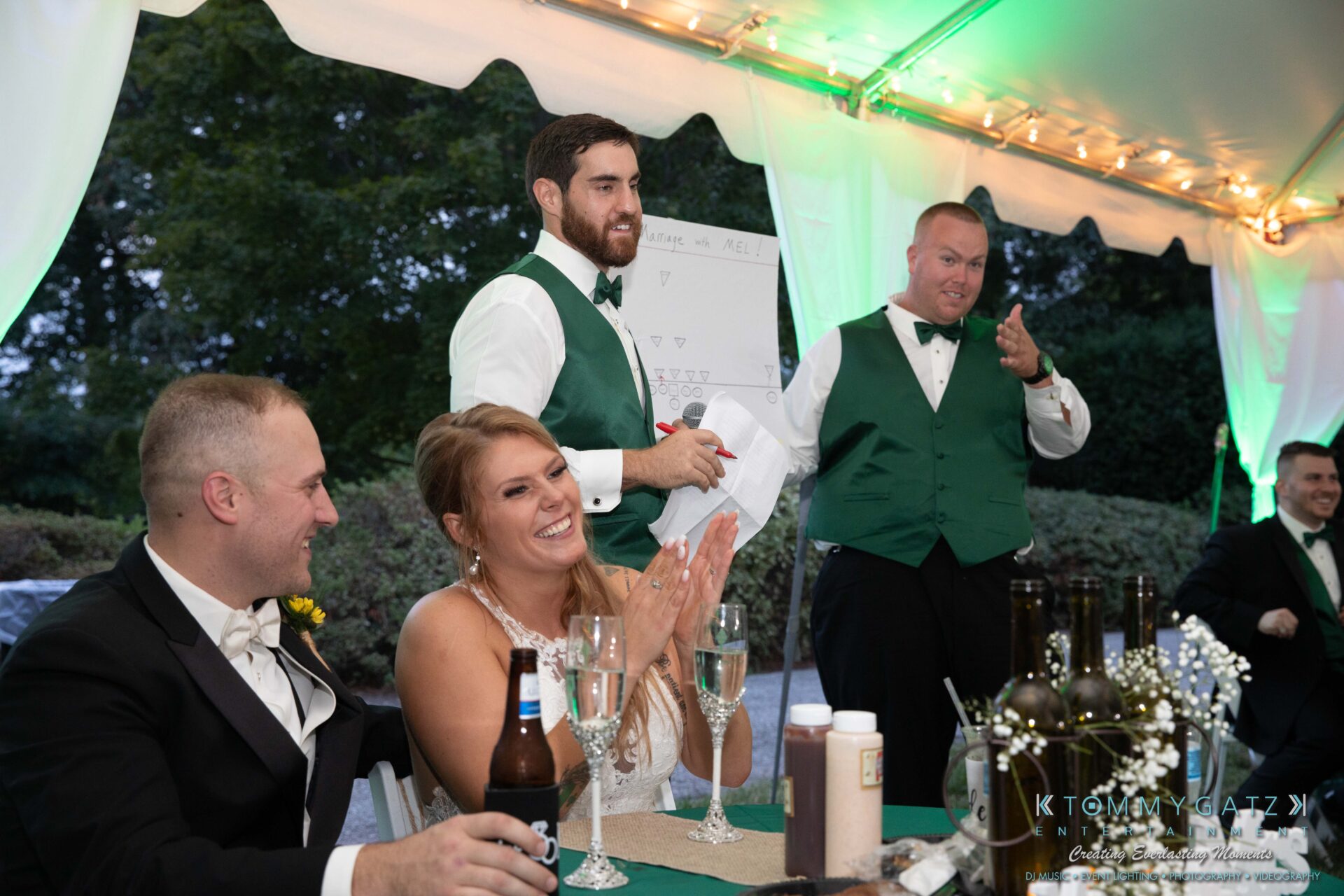 A bride and groom at a table with guests. One guest is standing, holding a microphone and speaking. The setting is an outdoor tented event with a board and bottles visible on the table.