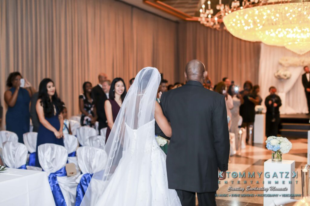Bride and man walking down the aisle at a wedding ceremony, guests standing and watching, chandelier overhead.