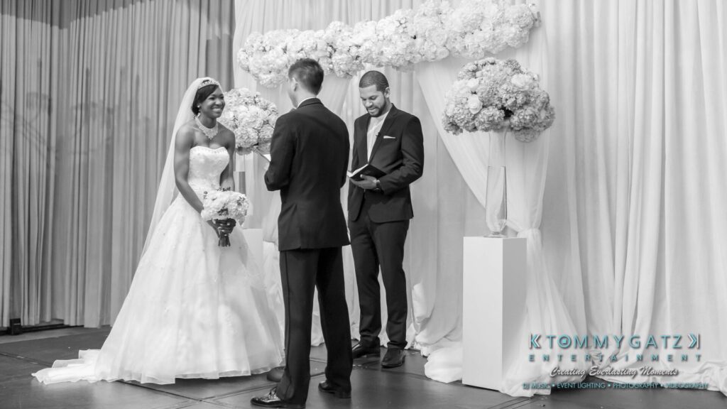 Bride and groom stand in front of an officiant during a wedding ceremony. The bride holds a bouquet, and decorative flower arrangements are visible in the background.