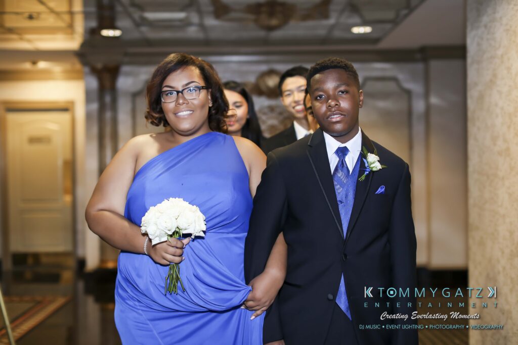 A woman in a purple gown holding white flowers walks with a man in a suit and bow tie. Others in formal attire follow behind them in a hallway.