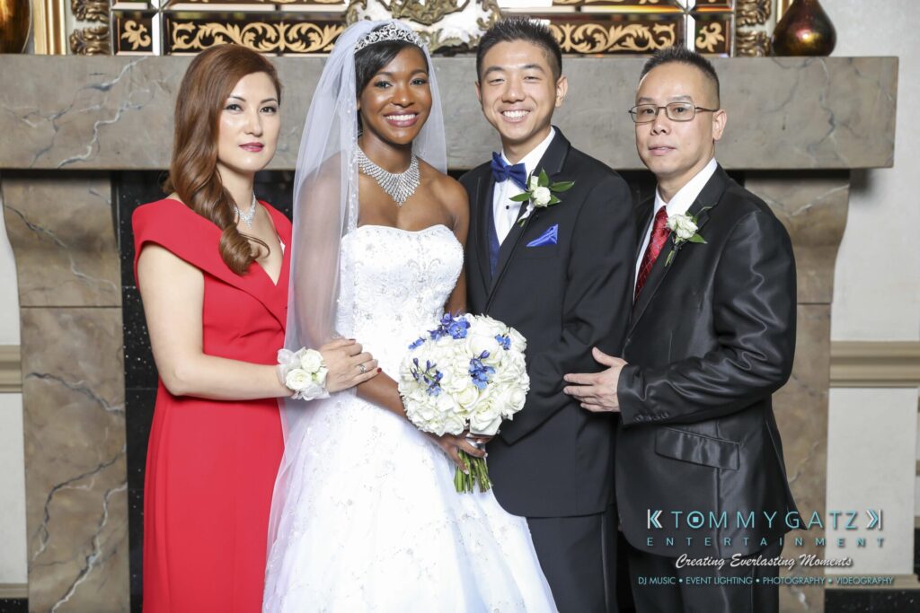 A wedding group photo with the bride in a white gown and veil, groom in a tuxedo, and two guests in formal attire standing beside them in front of a decorative fireplace.