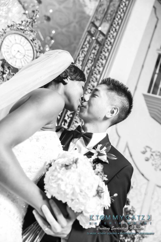 A bride and groom share a kiss in a black and white photo. The bride holds a bouquet, and both are dressed in formal wedding attire. Ornate decorations are visible in the background.