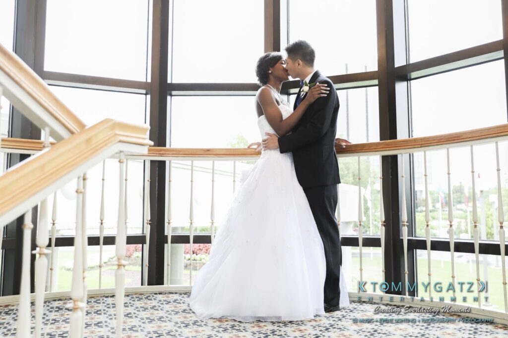 A couple in wedding attire shares a kiss on an indoor balcony with large windows in the background.