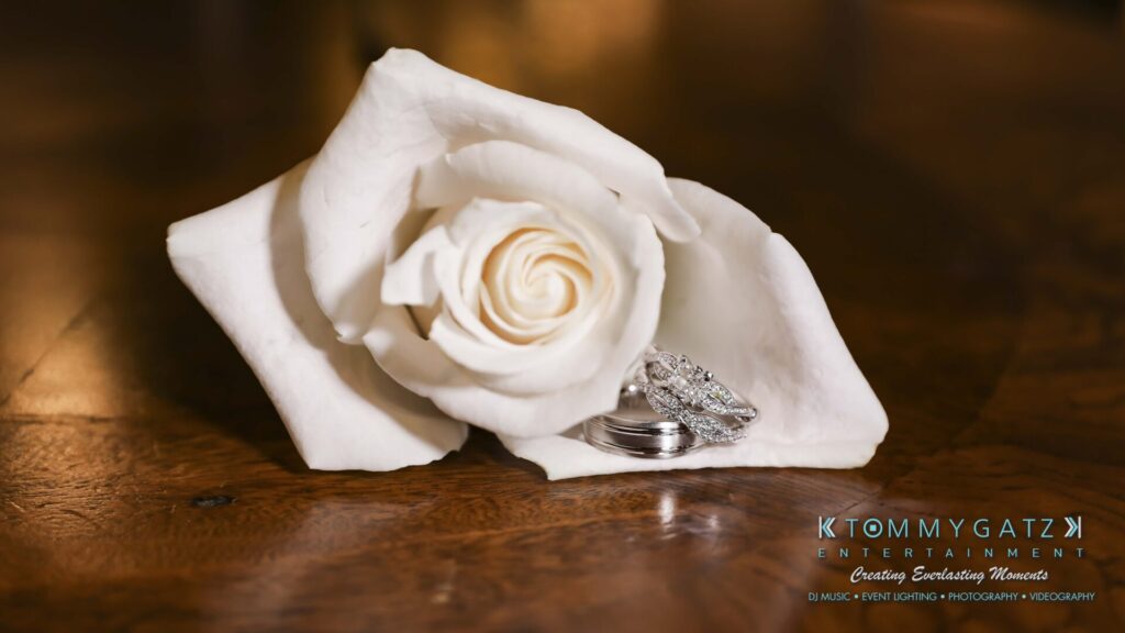 A white rose placed next to a set of wedding rings on a wooden surface, with a company logo in the lower right corner.