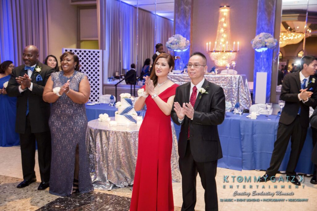 Guests in formal attire clap at an indoor event with decorated tables, silver tablecloths, and lit candles.
