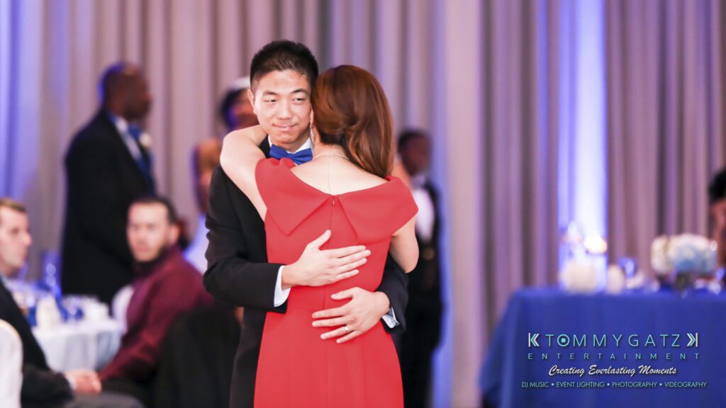 A couple dances closely in formal attire at an event. The background shows other attendees and a dining setup with blue lighting.