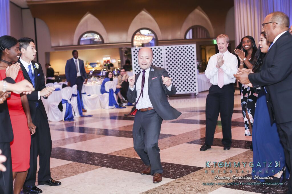 A man dances energetically in a formal setting while a group of people stands around watching and smiling.