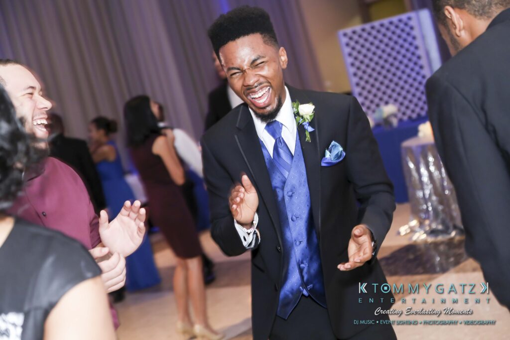 A man in a suit laughs joyfully at an indoor event, surrounded by others.