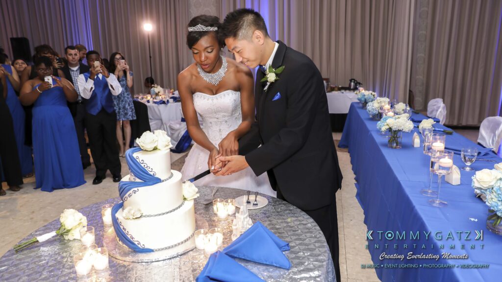 Bride and groom cutting a three-tiered wedding cake decorated with blue and white accents, surrounded by guests in a banquet hall.