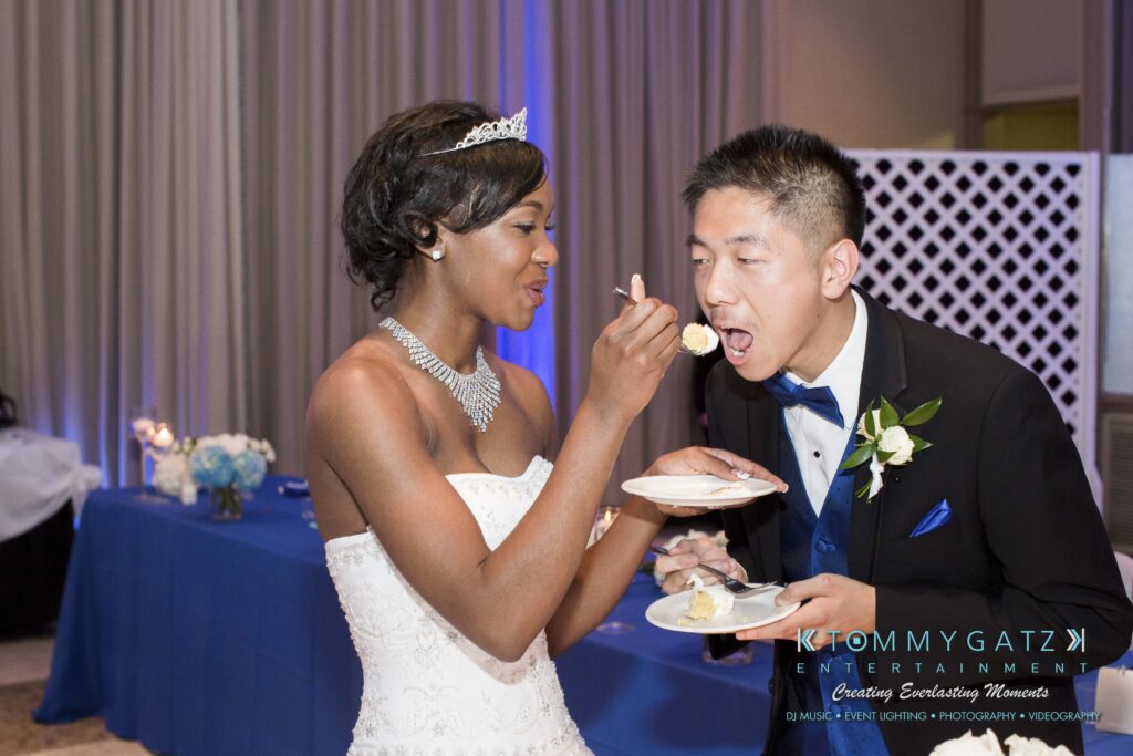 A bride in a white dress feeds cake to a groom in a tuxedo at a wedding reception.