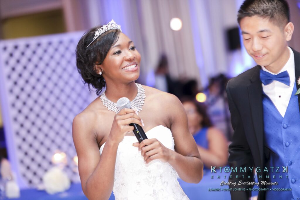 A person in a white dress with a tiara speaks into a microphone. Another person in a blue suit smiles beside them.