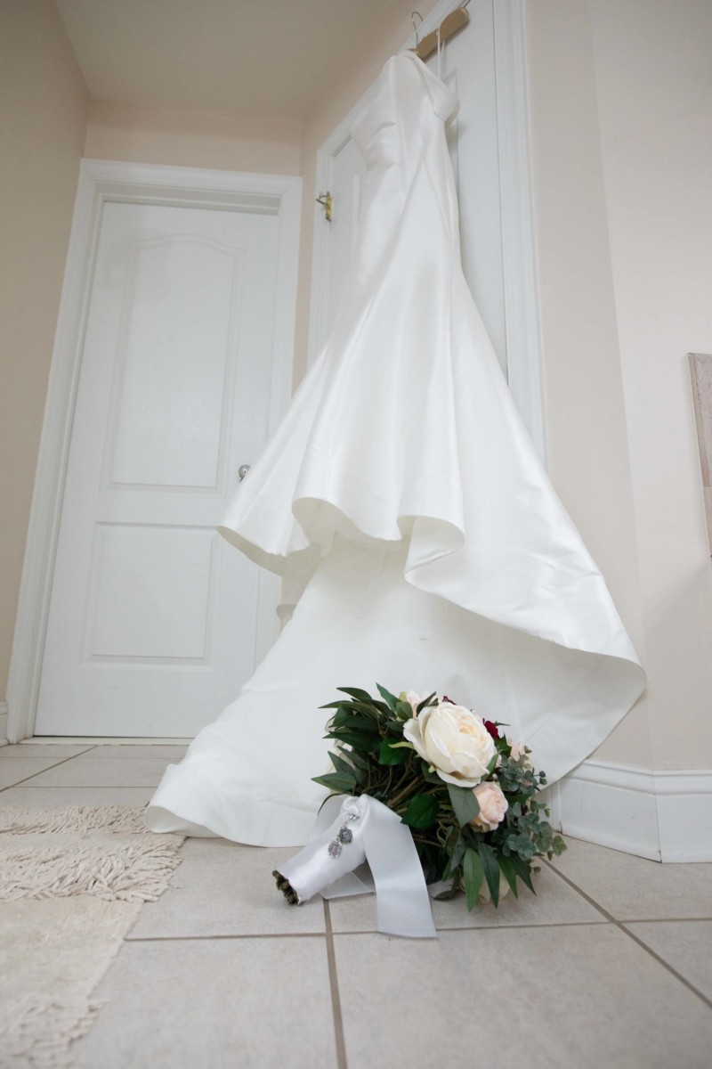 White wedding dress hanging on a door, with a bridal bouquet on the floor beneath it.