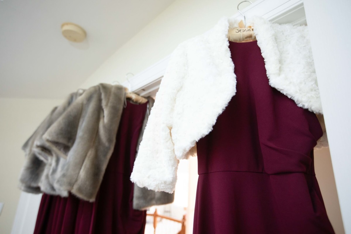 Three burgundy dresses hang on a white door, each topped with a faux fur shrug in gray, cream, and white.