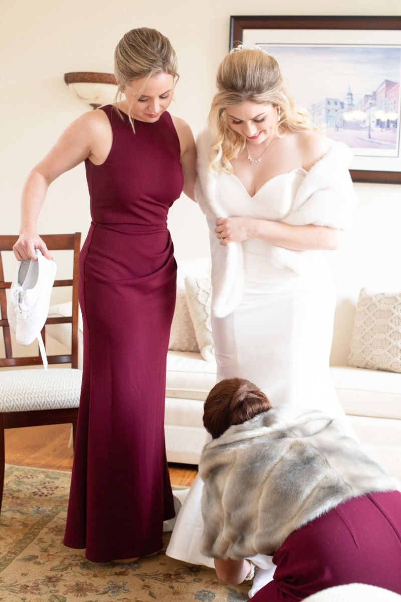 Two women assist a bride in a white gown with her train. One holds white shoes, and another crouches to adjust the dress. A painting hangs on the cream-colored wall behind them.