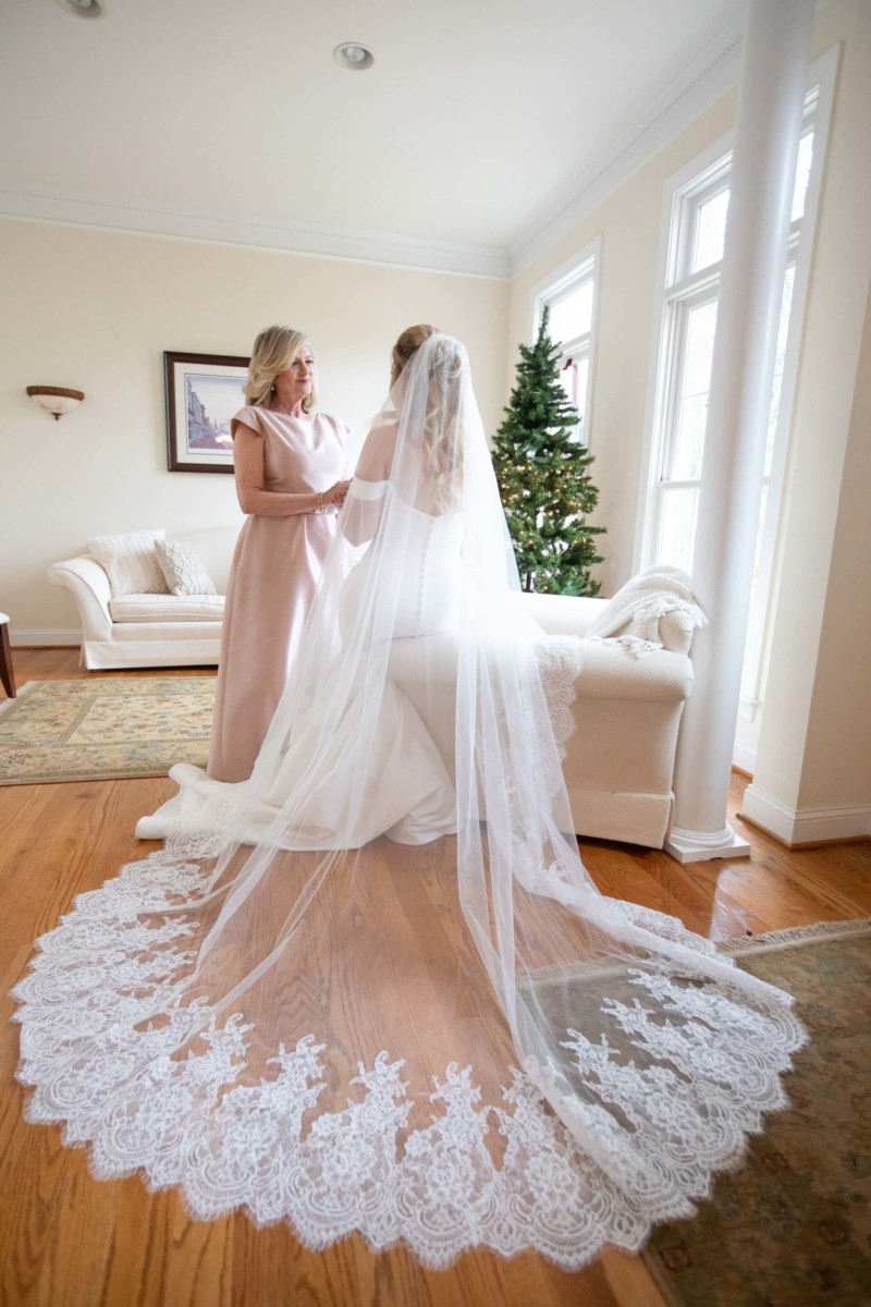 A bride in a white gown and veil sits with a woman in a pink dress in a living room. A decorated Christmas tree is visible in the background.