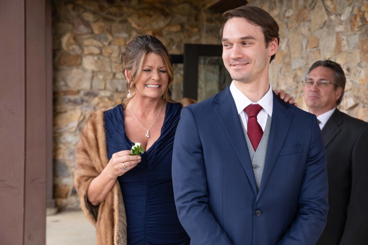 A woman stands smiling behind a man in a suit while holding a boutonniere. Another man is standing in the background. They are in front of a stone wall.
