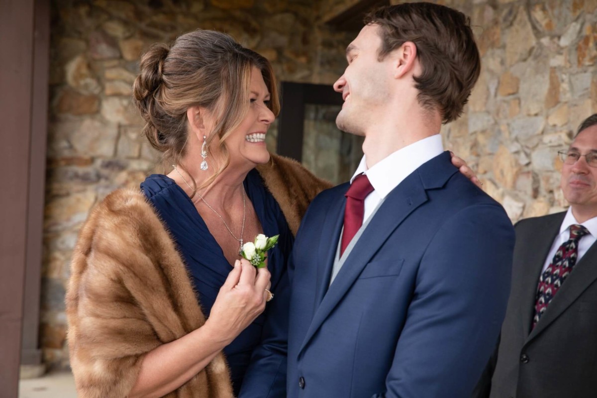 A woman in a fur stole laughs with a man in a blue suit. Another man in a suit stands nearby. They're outside near a stone wall.