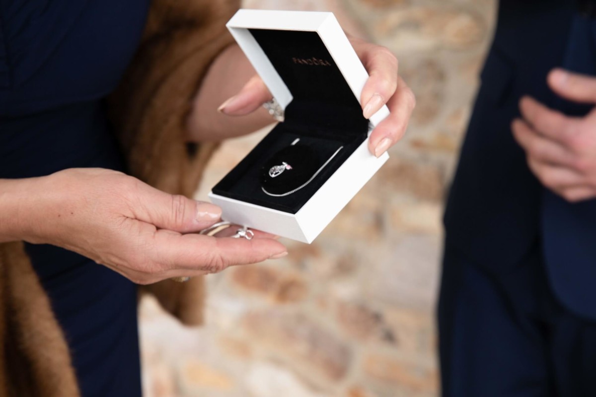 Person holding an open jewelry box displaying a necklace inside. Another person stands nearby.