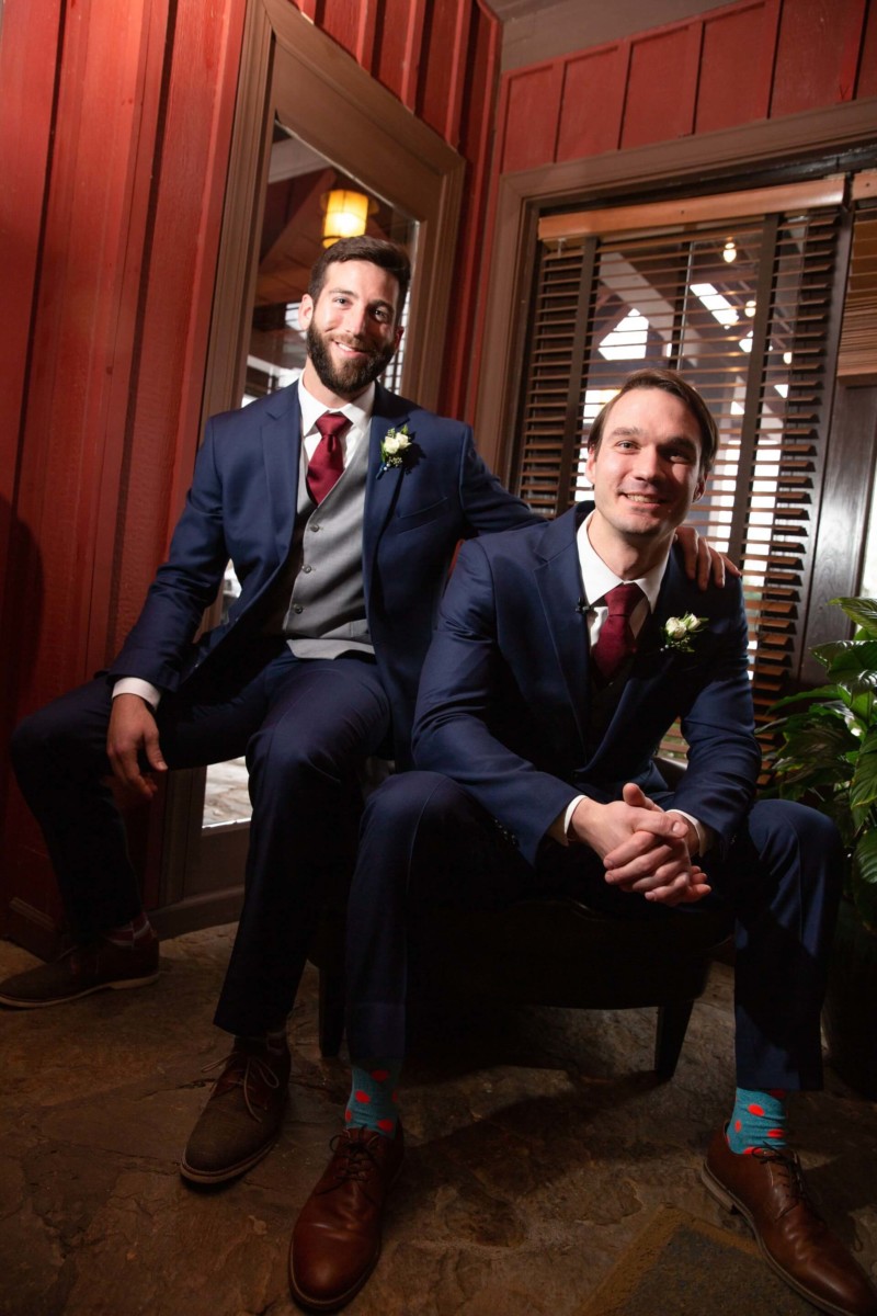Two men in suits, one seated and one resting his arm on the other's shoulder, pose indoors. They wear formal attire with boutonnieres.
