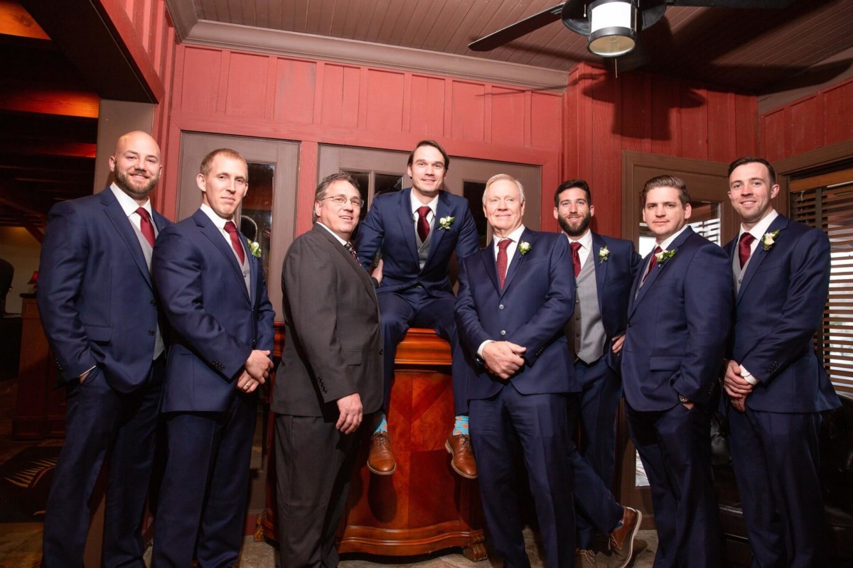 A group of eight men in formal wear, including blue suits and ties, pose in a room with wood-paneled walls. One man sits on a bar counter while the others stand around him.