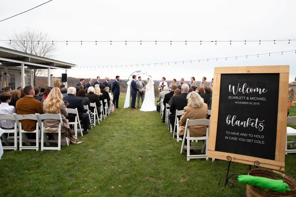 Outdoor wedding ceremony with guests seated on white chairs, a couple at the altar, and a sign offering blankets.