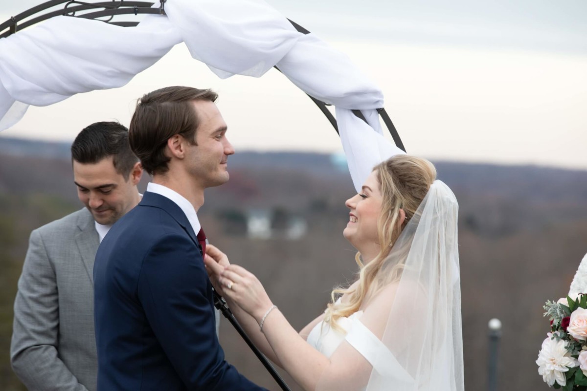A couple stands under an arch during their wedding ceremony, with a person in a suit nearby. The bride adjusts the groom's attire.