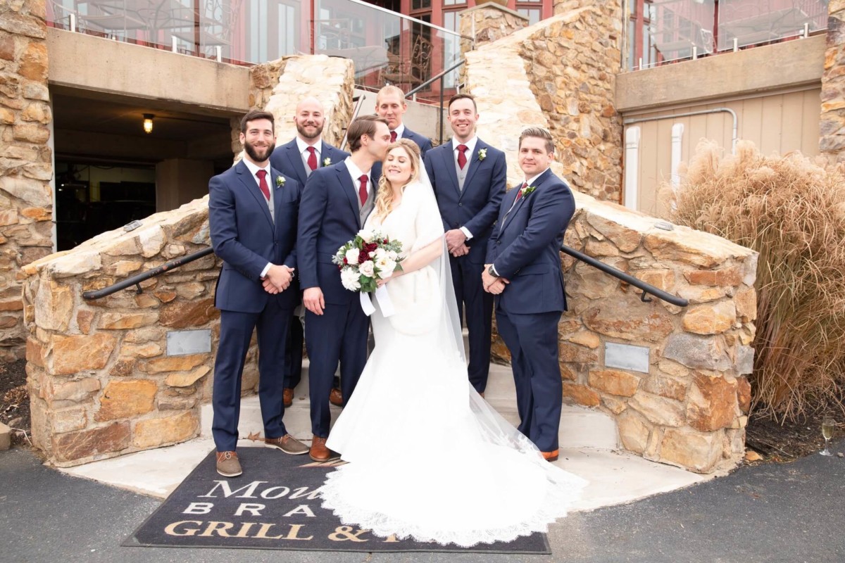A bride and six groomsmen in formal attire pose on stone steps outside a building.