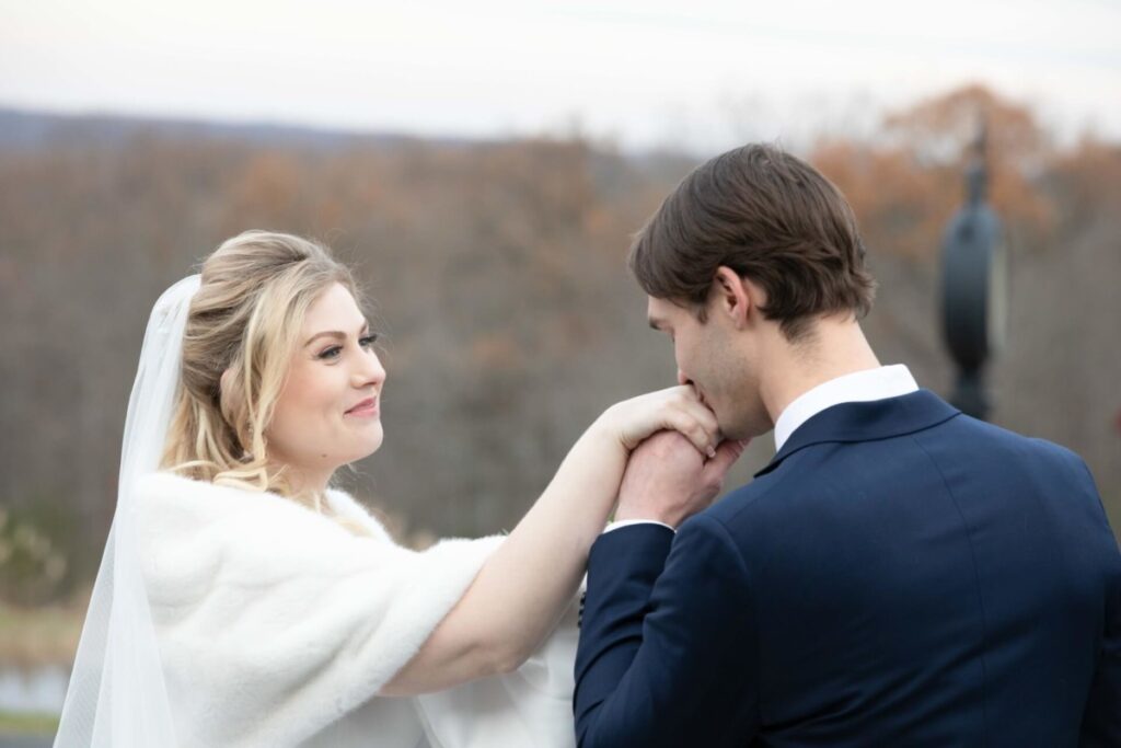 A bride in a white dress smiles as the groom in a dark suit kisses her hand outdoors.