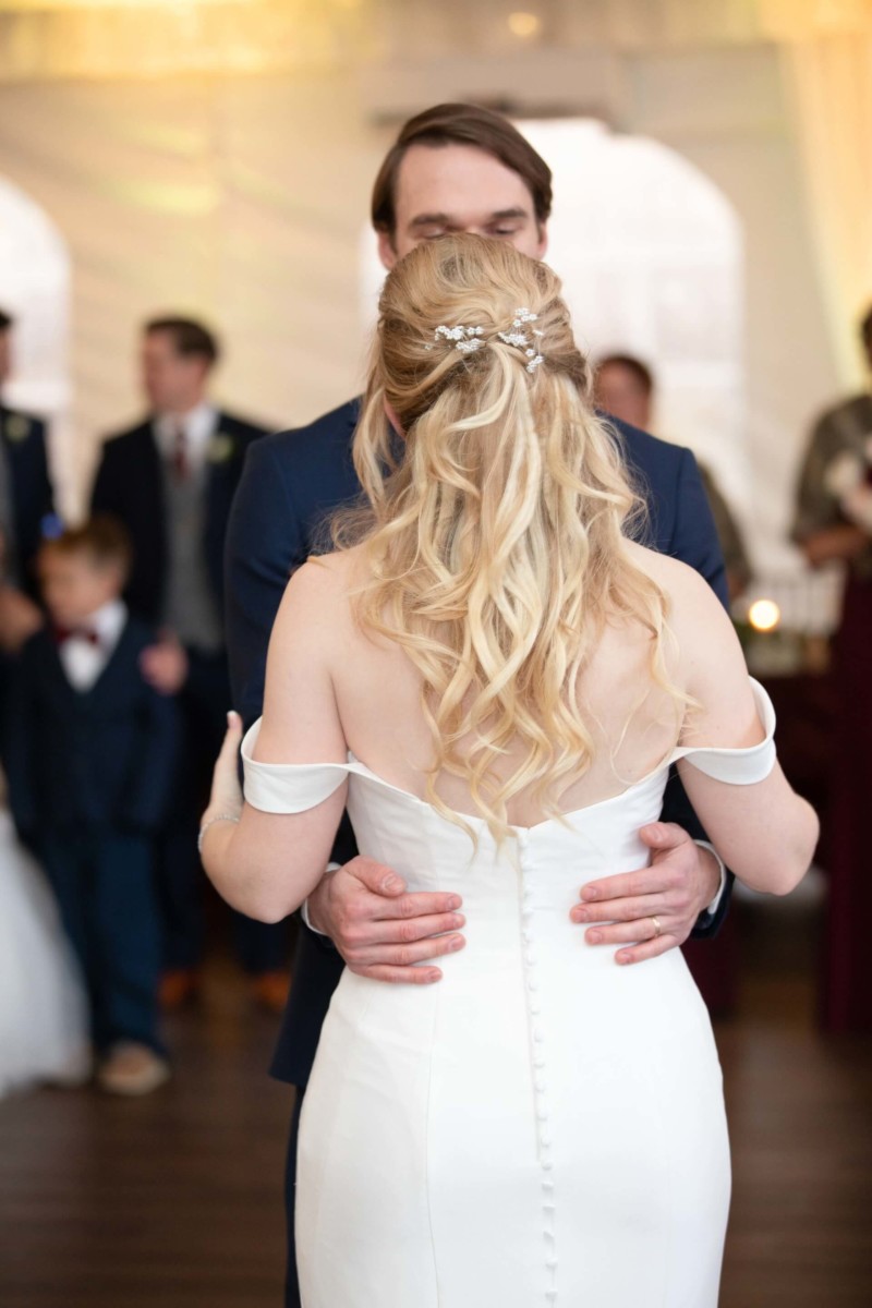 A couple shares a dance at a wedding, with the bride in a white dress and the groom in a dark suit. Guests are blurred in the background.