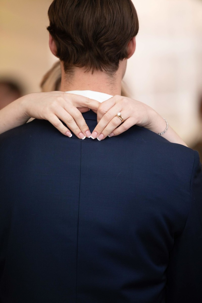 A person with short hair is seen from behind, wearing a dark suit. Two hands with a ring and bracelet are gently resting on the person's shoulders.