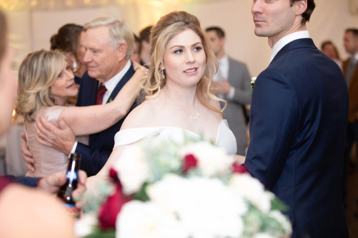 A bride and groom stand together at a reception. The bride holds a bouquet with red and white flowers. Guests are seen in the background, with some dancing and embracing.