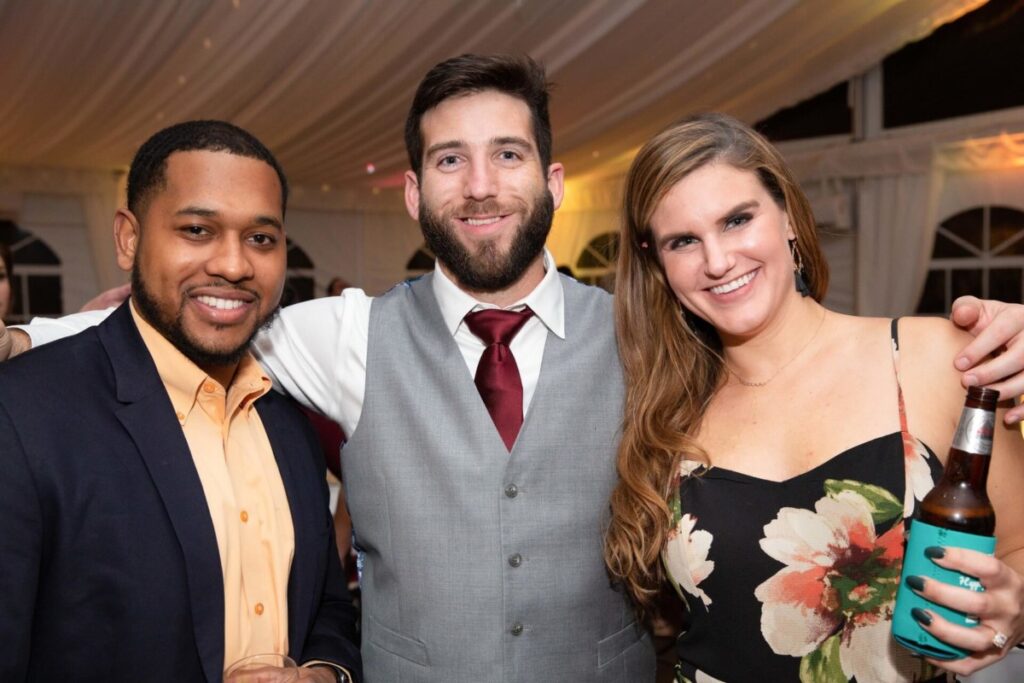 Three people standing together at an event, smiling at the camera. One holds a drink bottle, and another holds a glass. They're dressed in semi-formal attire.