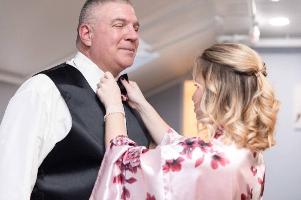 A woman adjusts a man's bow tie. The man wears a white shirt with a black vest, and the woman is in a floral satin robe. They are indoors with ceiling lights in the background.