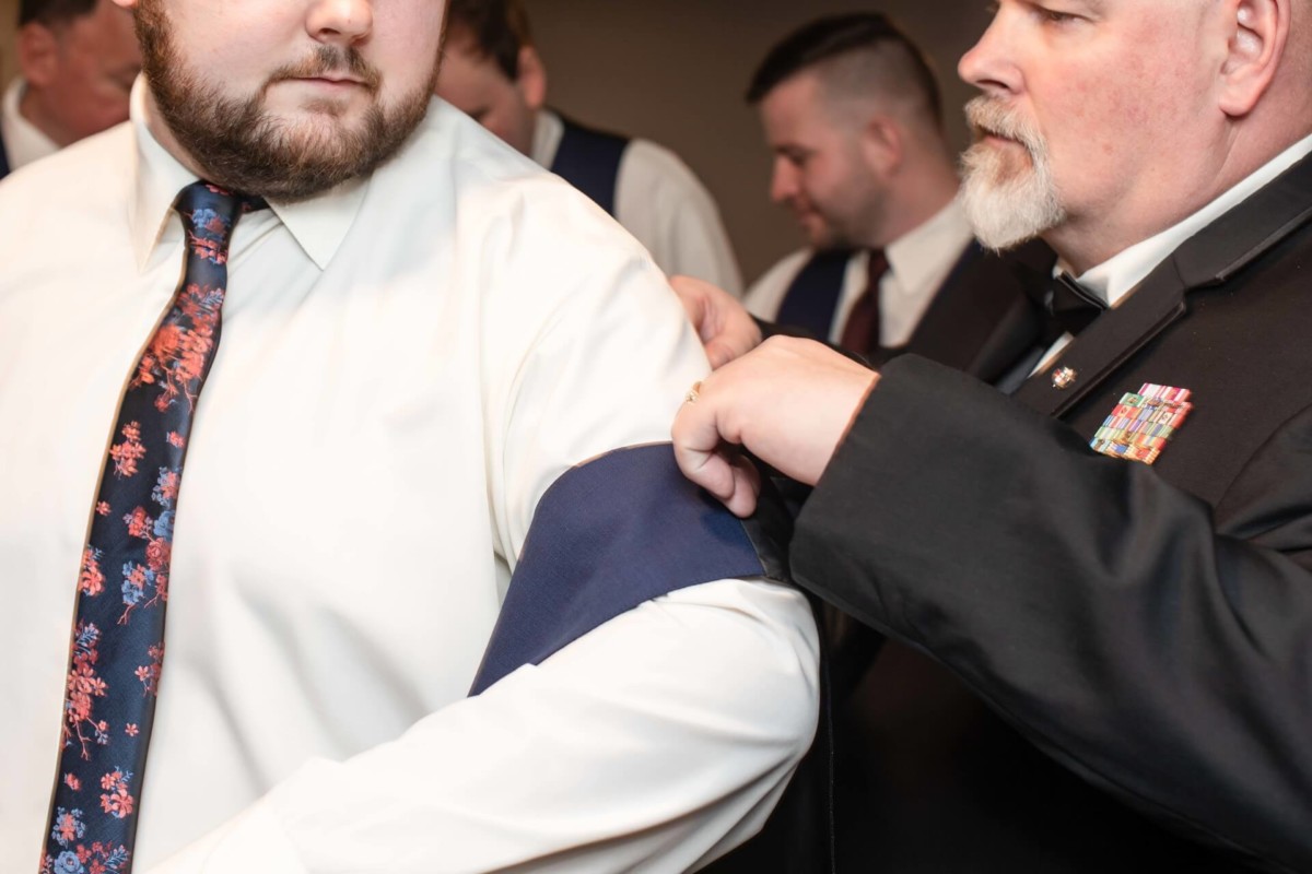 A man helps another man put on a blue sash. Both are wearing formal attire, and there are other men in the background, also in formal clothing.