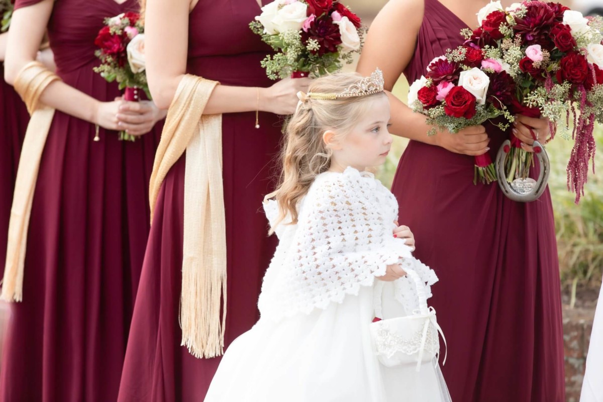 A young girl in a white dress stands in front of women wearing burgundy gowns and holding bouquets of red and white flowers.