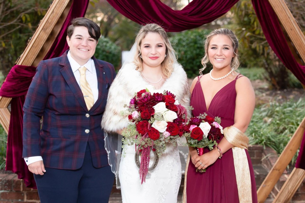 Three people dressed in formal attire stand outdoors. The center person holds a bouquet of red and white flowers. They are framed by a wooden arch draped with burgundy fabric.