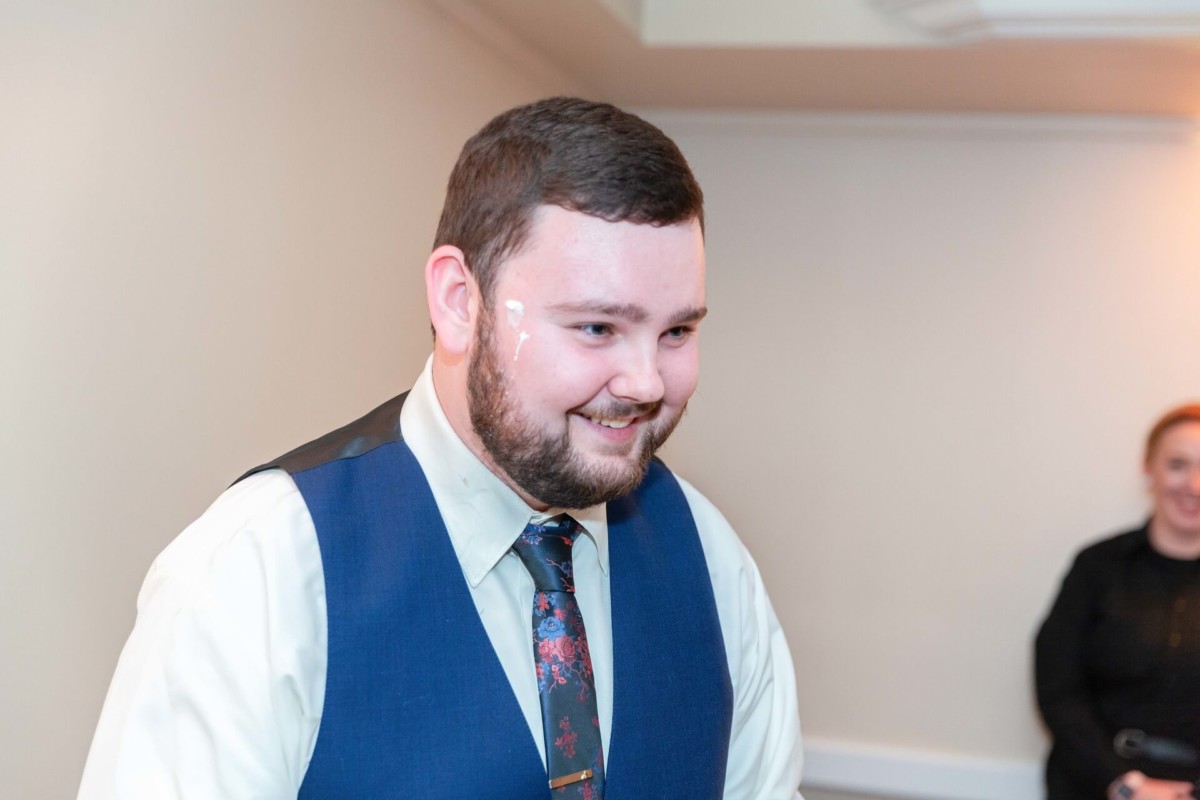 A man with a beard smiling, wearing a blue vest and tie, stands in a room with white walls. A woman in black is visible in the background.