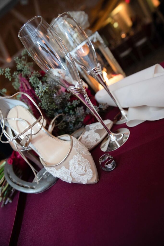 Wedding still life with lace high-heeled shoes, two champagne flutes, and rings on a burgundy tablecloth.