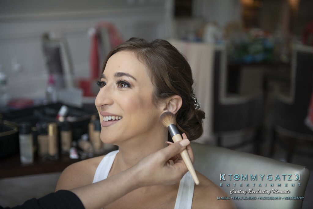 Person having makeup applied by another person, seated in a room with makeup products nearby.