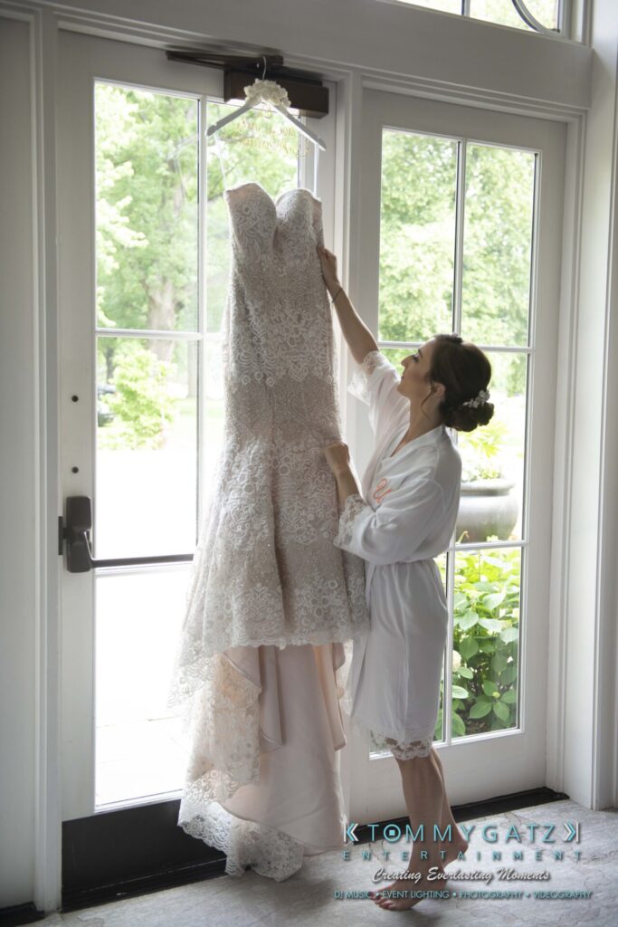 A woman in a robe admires a lace wedding dress hanging by a window.