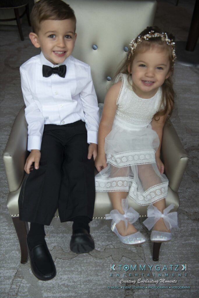 Two young children dressed in formal attire, sitting on a chair. The boy is in a white shirt and bow tie, and the girl is in a white dress with decorative bows on her shoes.