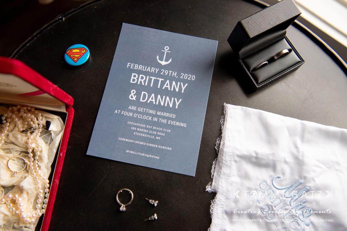 Invitation for Brittany and Danny's wedding on February 29, 2020, next to jewelry, a Superman pin, a pocket watch, and a handkerchief on a table.